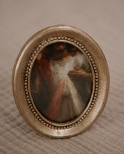Catholic home decor in a small oval painting of the divine mercy image in a decorative gold frame on a textured beige surface