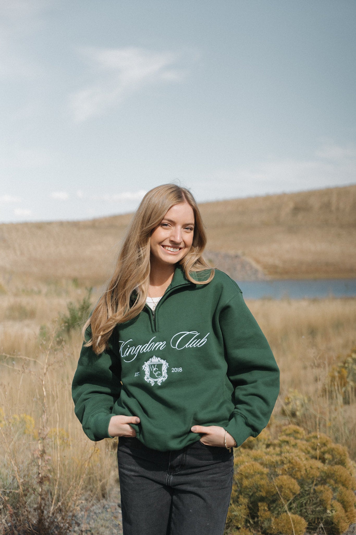Woman wearing a green 'Kingdom Club' sweatshirt in a field with a lake in the background