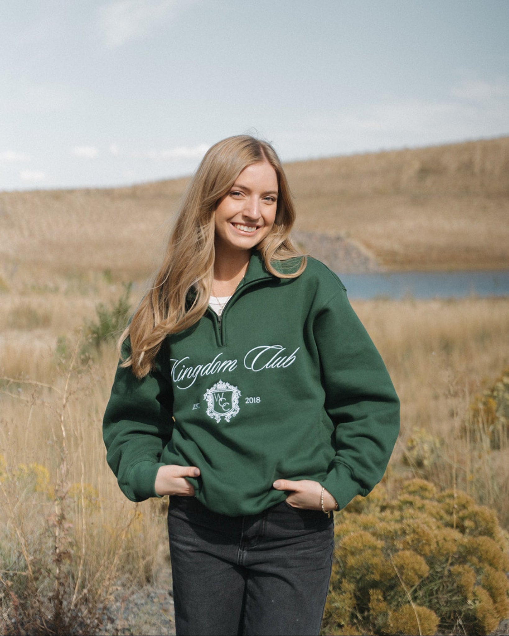Woman wearing a green 'Kingdom Club' sweatshirt in a field with a lake in the background