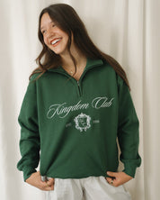 Woman wearing a green 'Kingdom Club' sweatshirt against a white curtain background