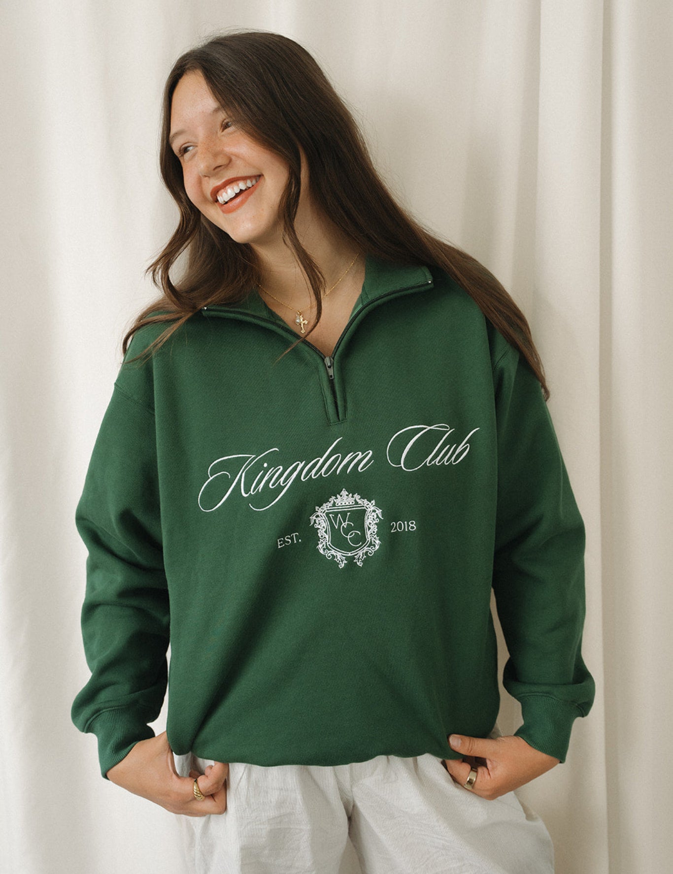 Woman wearing a green 'Kingdom Club' sweatshirt against a white curtain background