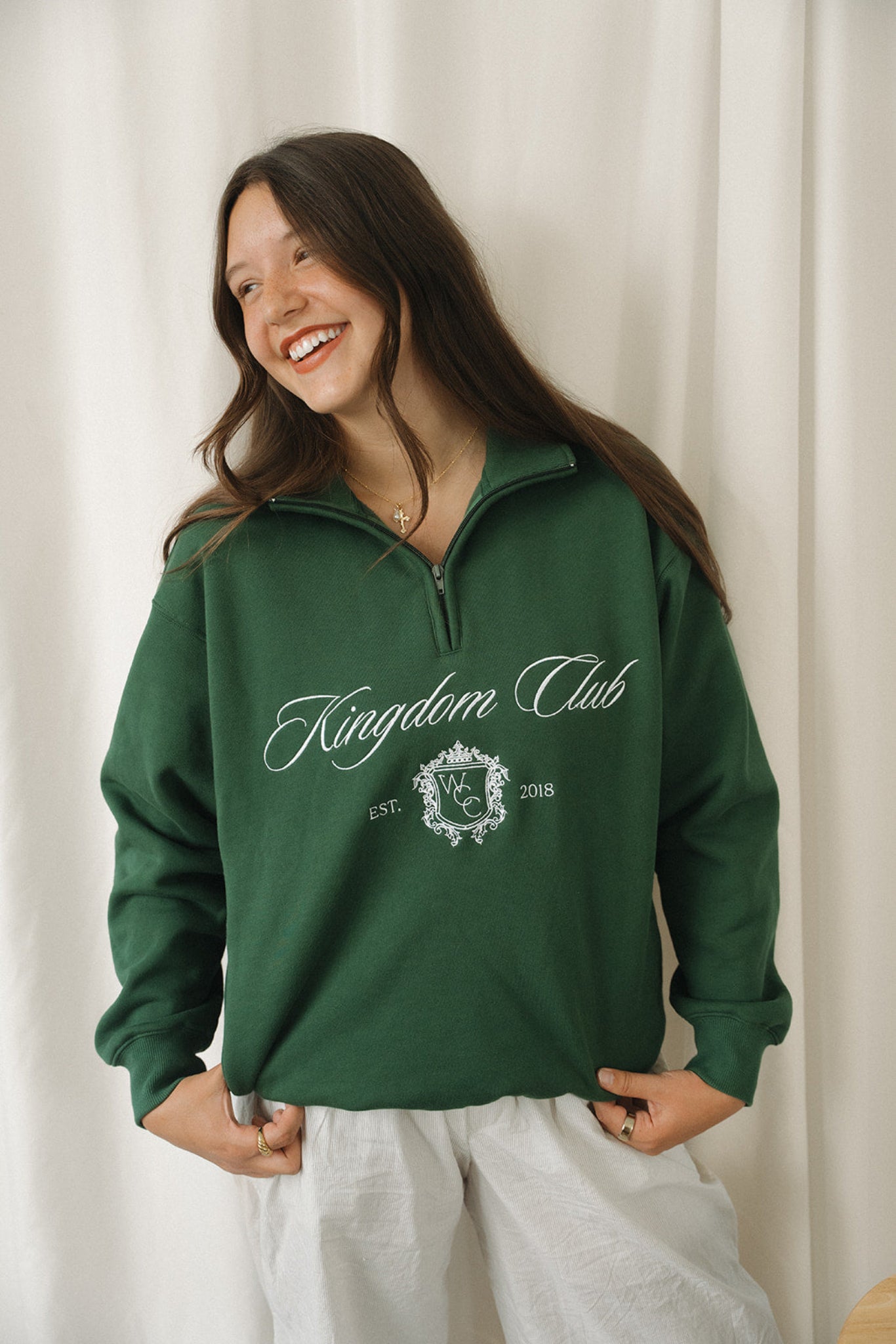 Woman wearing a green 'Kingdom Club' sweatshirt against a white curtain background