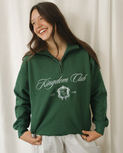 Woman wearing a green 'Kingdom Club' sweatshirt against a white curtain background