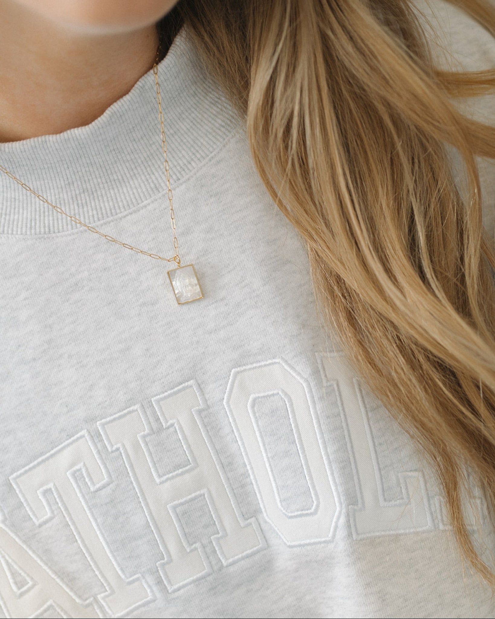Person wearing a gray sweatshirt with 'CATHOLIC' text and a gold necklace with a square pendant.