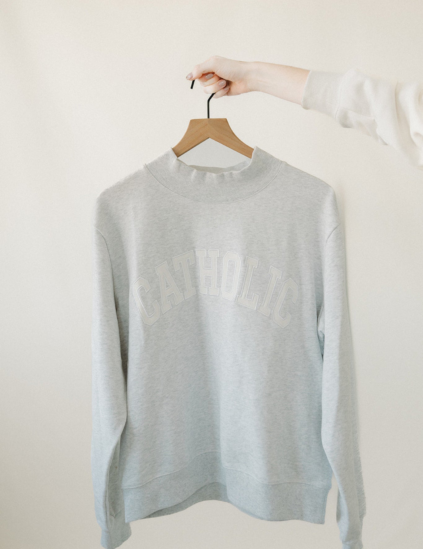 Light gray catholic varsity sweater on a hanger against a white background