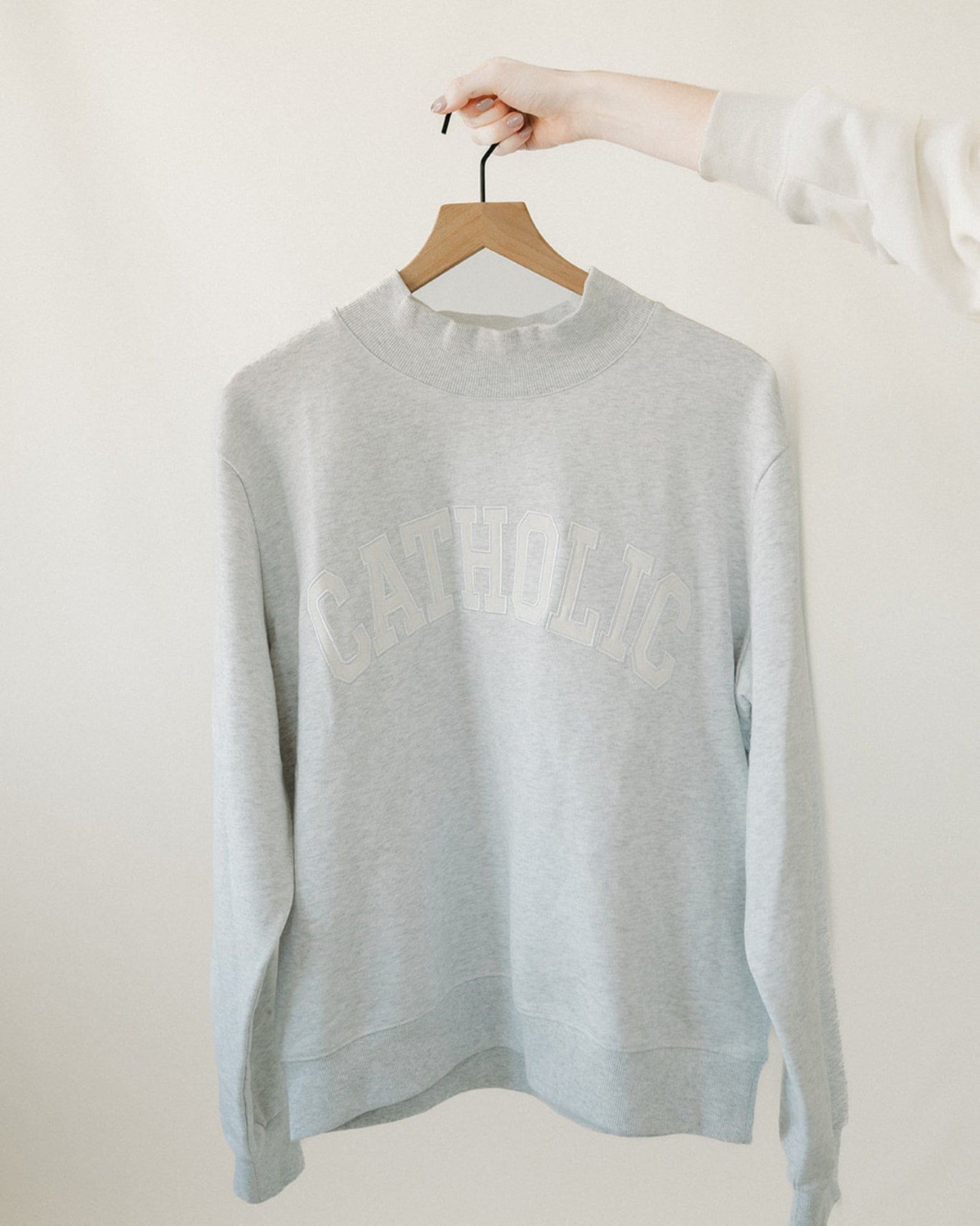 Light gray catholic varsity sweater on a hanger against a white background