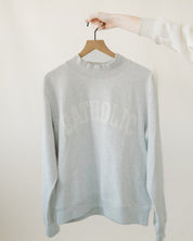 Light gray catholic varsity sweater on a hanger against a white background