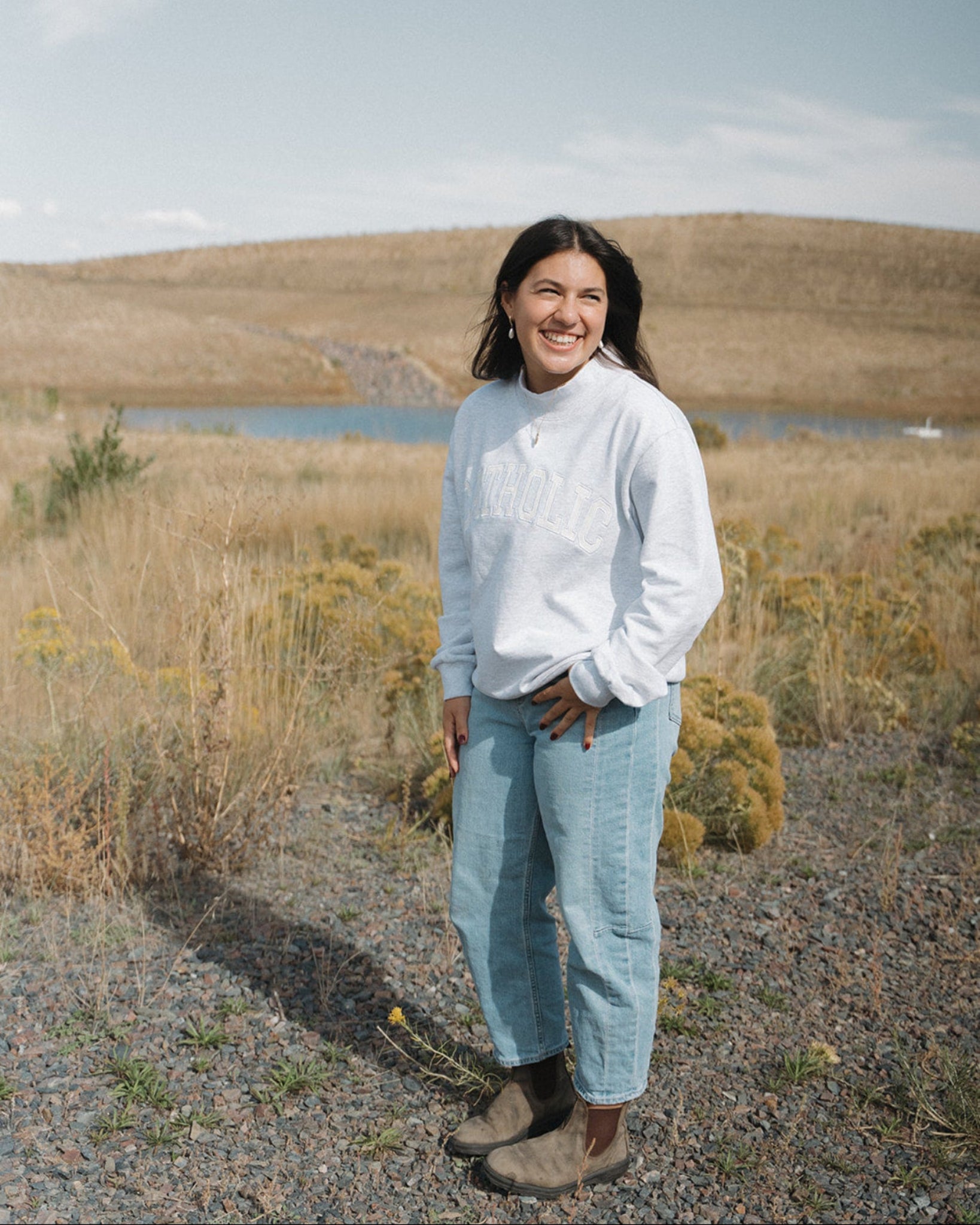 Woman standing in a field wearing a West Coast Catholic varsity mock-neck crewneck, jeans, and blunt stones. 