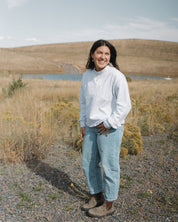 Woman standing in a field wearing a West Coast Catholic varsity mock-neck crewneck, jeans, and blunt stones. 