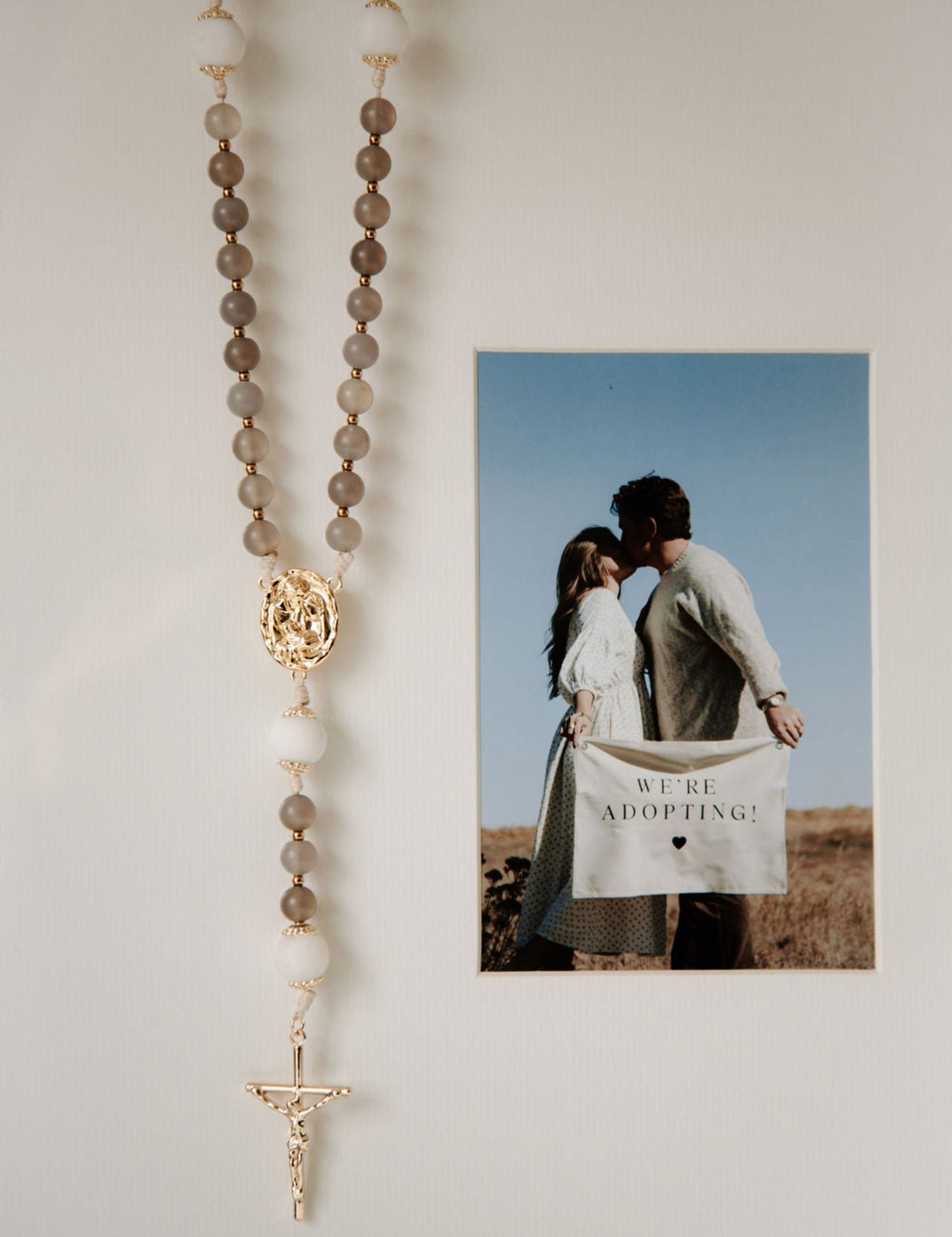 Rosary hanging on a wall next to a photo of a couple holding a 'We Are Adopting!' card.