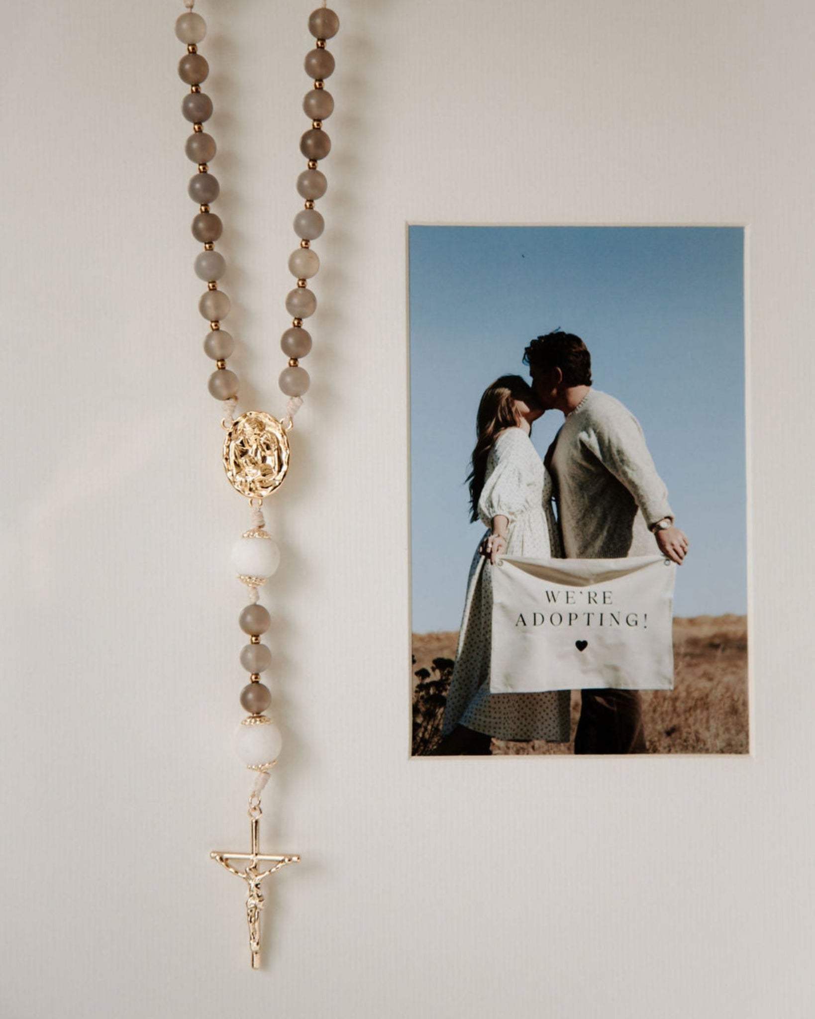 Rosary hanging on a wall next to a photo of a couple holding a 'We Are Adopting!' card.