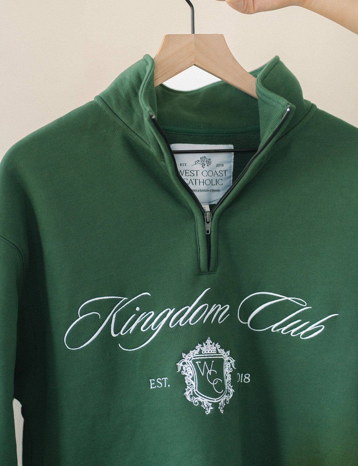 Green sweatshirt with 'Kingdom Club' text and logo on a hanger against a beige background