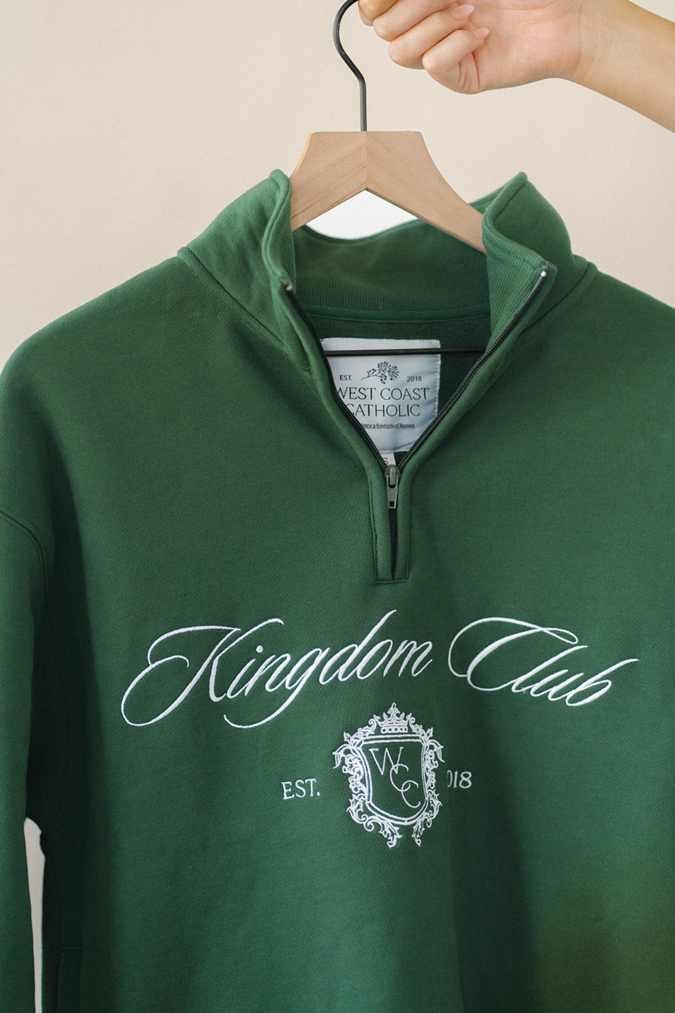 Green sweatshirt with 'Kingdom Club' text and logo on a hanger against a beige background