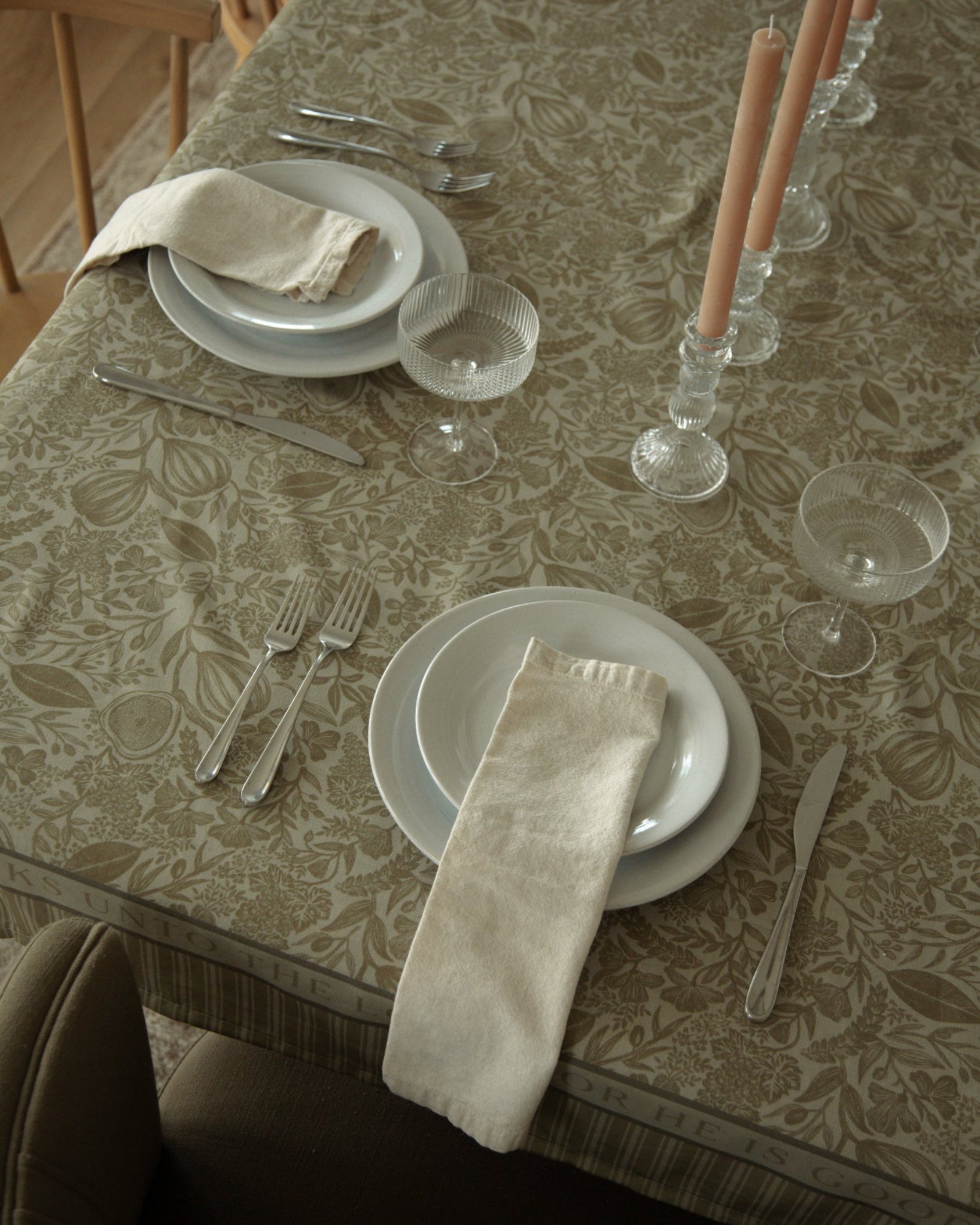 A styled dining table setting featuring a beige and brown floral patterned Catholic tablecloth, white plates, folded cream napkins, clear glass goblets, and candlesticks with light pink taper candles.