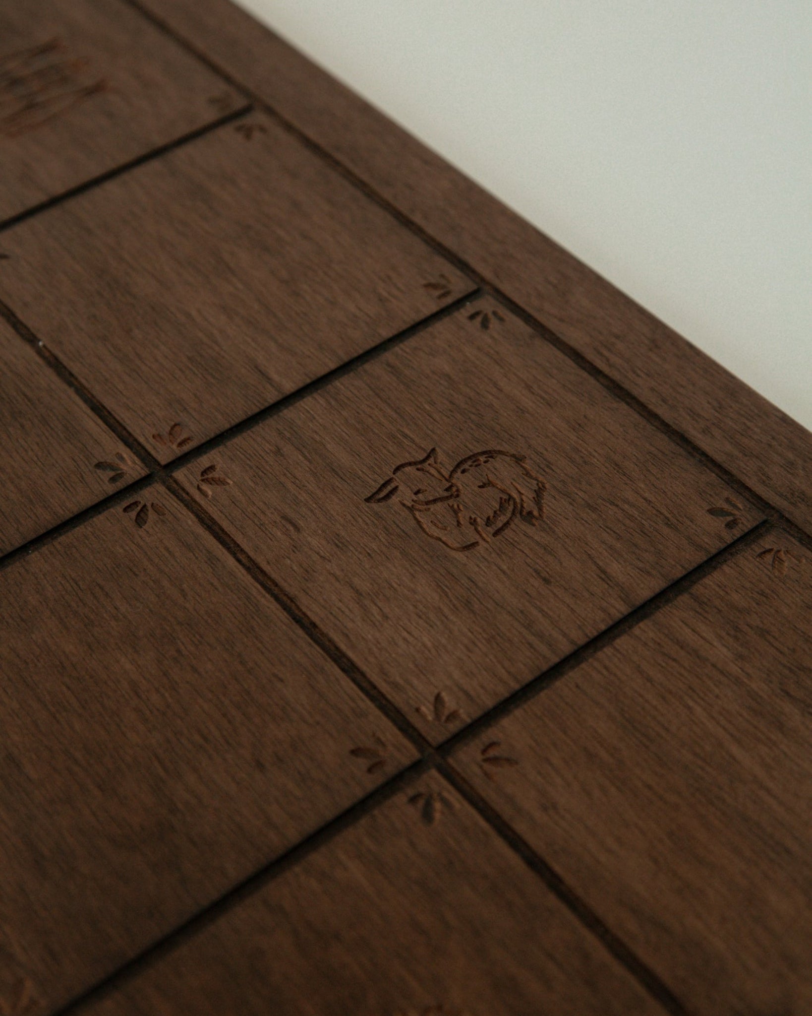 A close-up of a Catholic dark brown wooden board with engraved square sections and a small carved animal emblem in the center, photographed against a light neutral background.