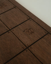 A close-up of a Catholic dark brown wooden board with engraved square sections and a small carved animal emblem in the center, photographed against a light neutral background.