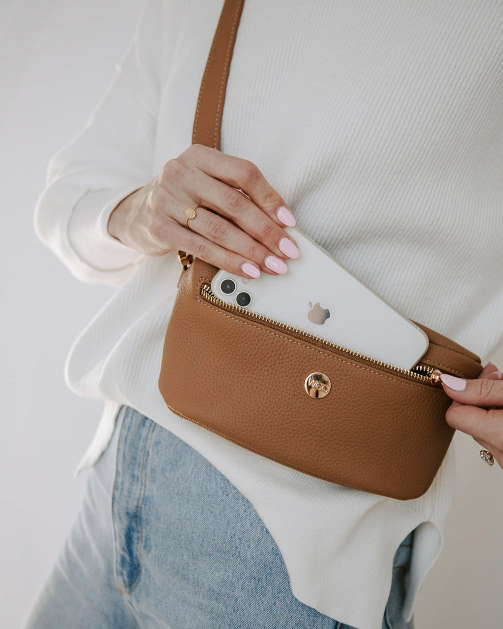 Person holding a brown crossbody bag with a white phone case inside, wearing a white sweater and blue jeans.