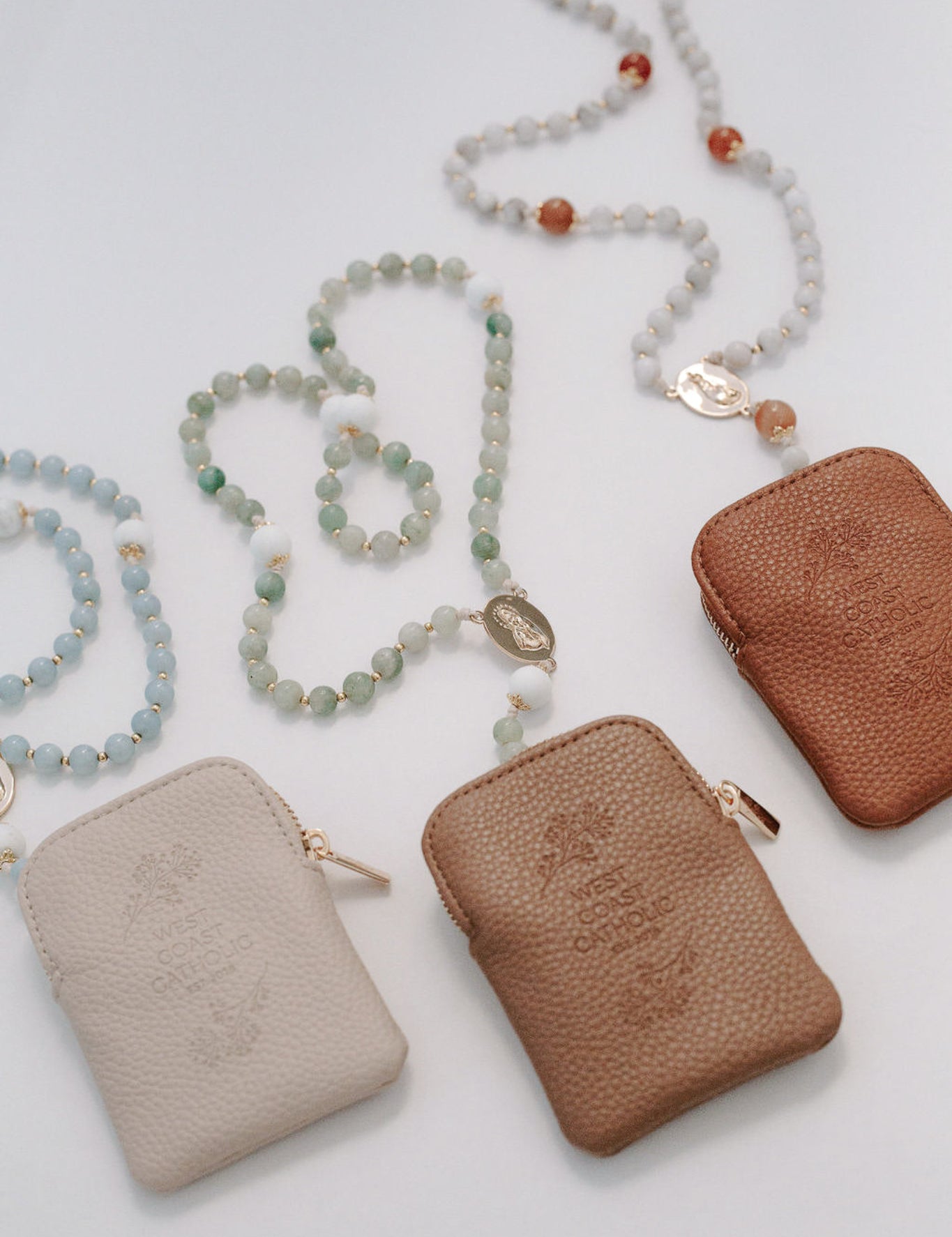 Three leather pouches in beige, brown, and brown with rosaries on a light gray background