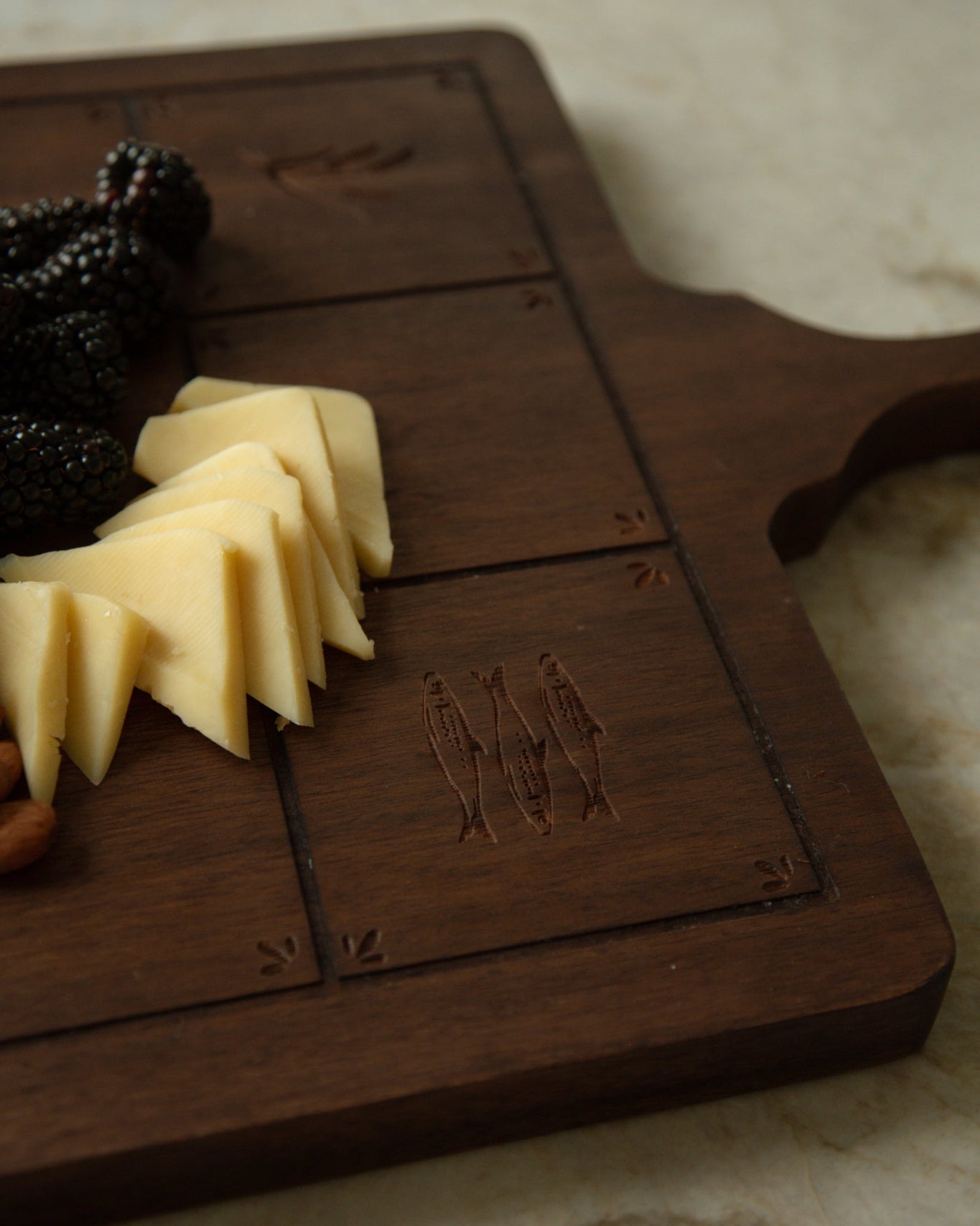A dark brown wooden serving board with carved fish Catholic Imagery, styled with cheese wedges, blackberries, and a nut on a light marble surface.