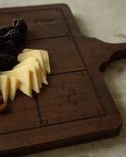A dark brown wooden serving board with carved fish Catholic Imagery, styled with cheese wedges, blackberries, and a nut on a light marble surface.