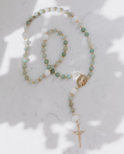 A rosary with green and blue-green beads and gold accents, displayed on a white surface with a shadow cast over it.