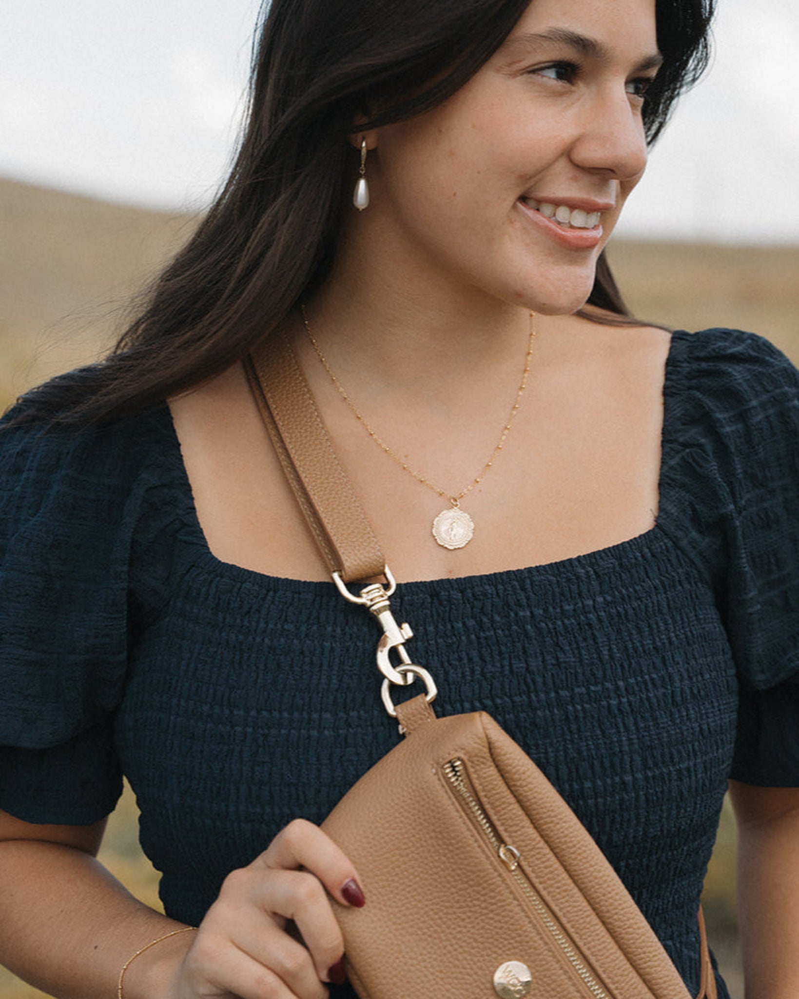 Woman holding a brown leather bag, wearing a miraculous medal necklace, outdoors with a blurred natural background