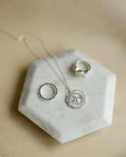 Sterling silver jewelry set including a round miraculous medal necklace and catholic rings, displayed on a white hexagonal marble tray over a light wood surface.