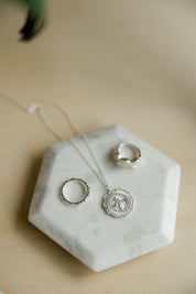 Sterling silver jewelry set including a round miraculous medal necklace and catholic rings, displayed on a white hexagonal marble tray over a light wood surface.