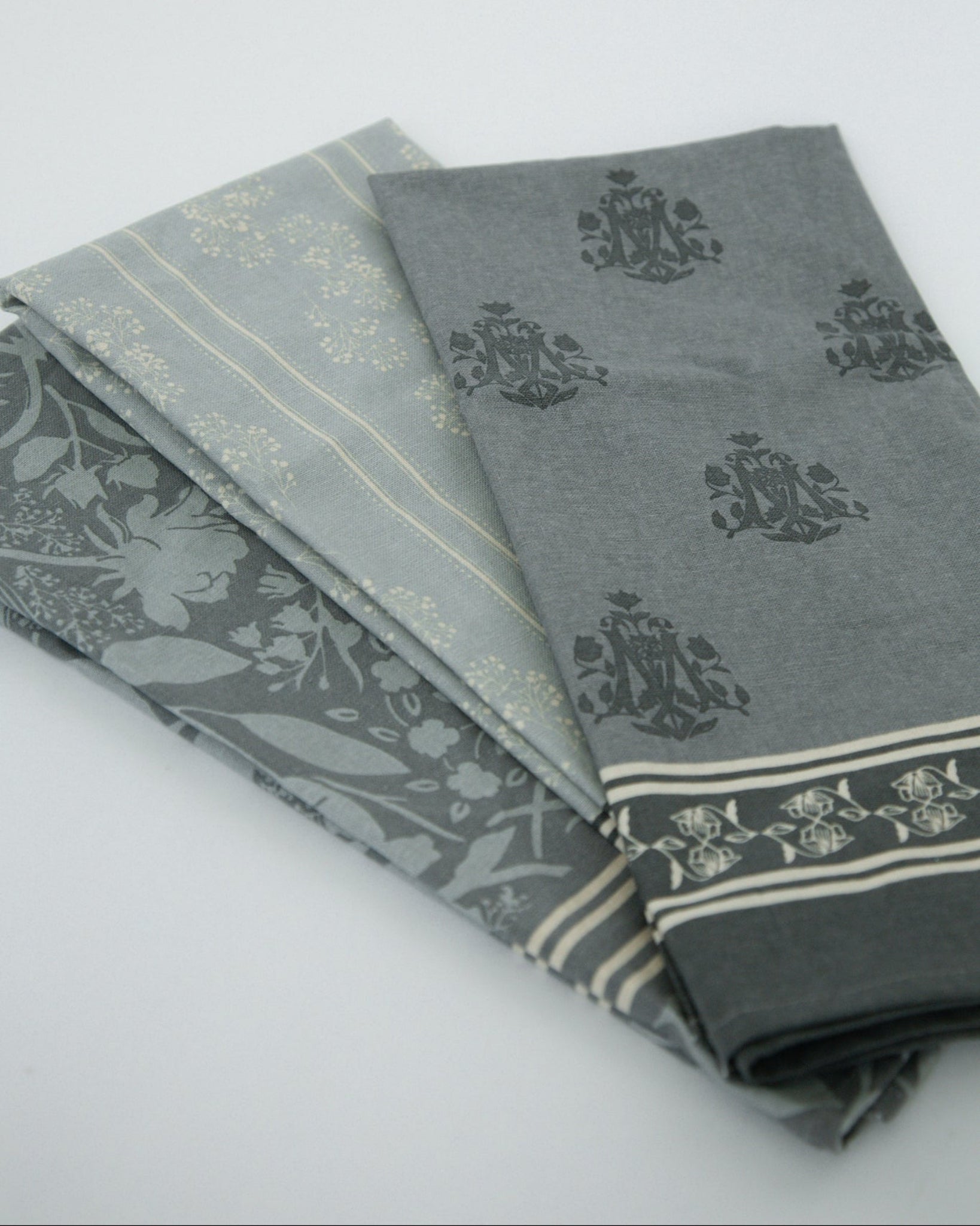 Three folded Marian inspired tea towels in shades of blue/gray with floral and striped patterns, arranged on a light gray background.