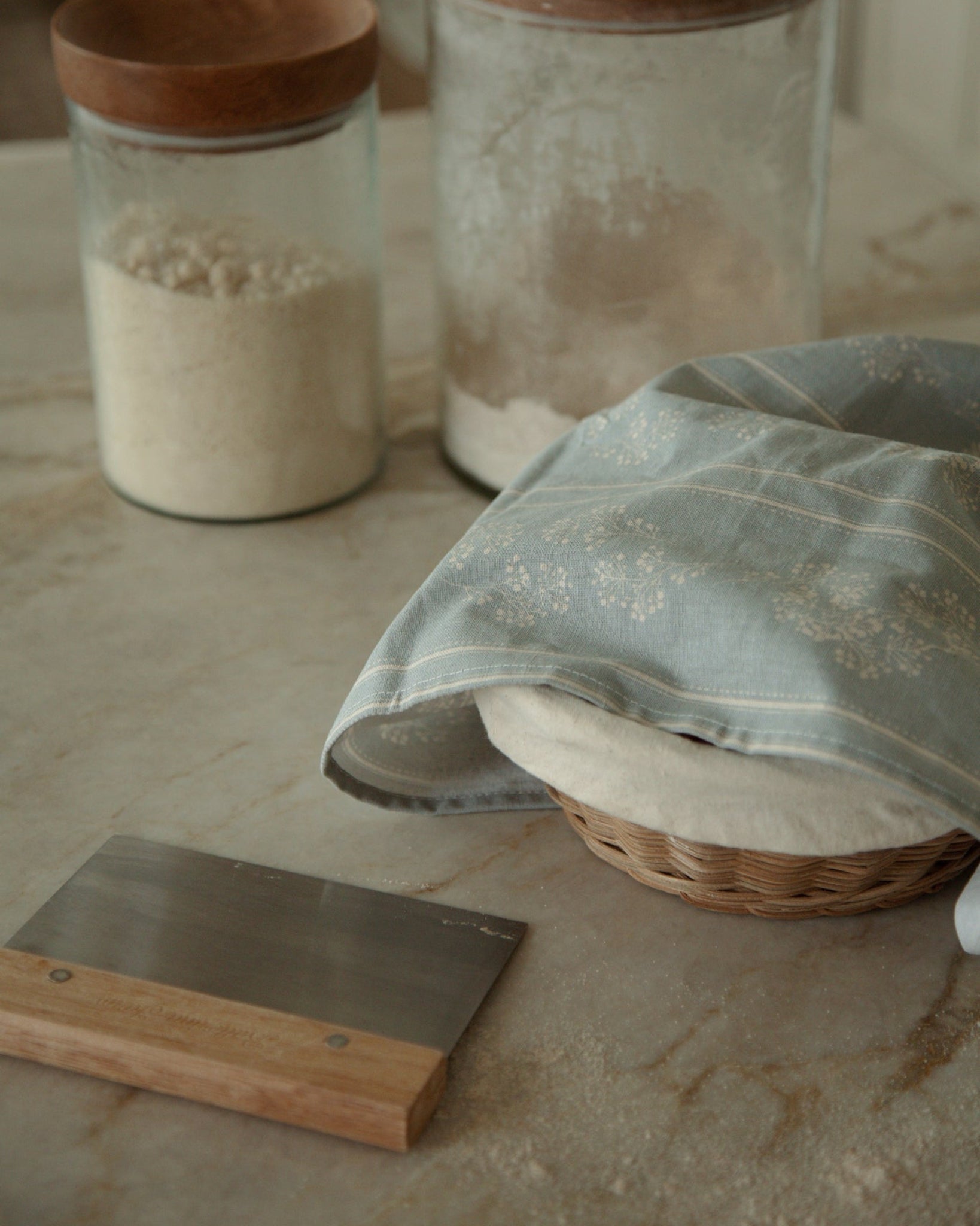 Two clear glass canisters with round wooden lids, one filled with flour and the other with sugar, set on a stone countertop beside a blue babies breath patterned kitchen towel, a small wicker basket, and a wooden-and-metal scraper.