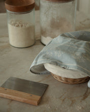 Two clear glass canisters with round wooden lids, one filled with flour and the other with sugar, set on a stone countertop beside a blue babies breath patterned kitchen towel, a small wicker basket, and a wooden-and-metal scraper.