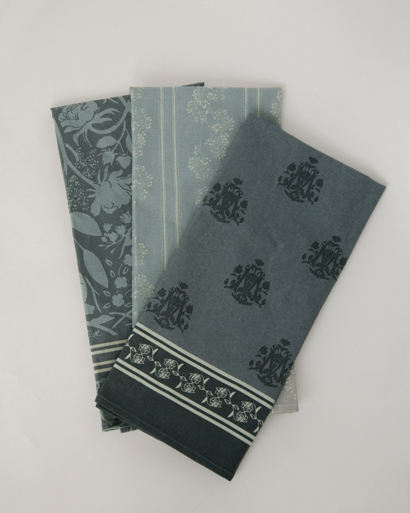 Three folded cotton towels in muted blue-green tones with floral and striped patterns, arranged on a light neutral background.