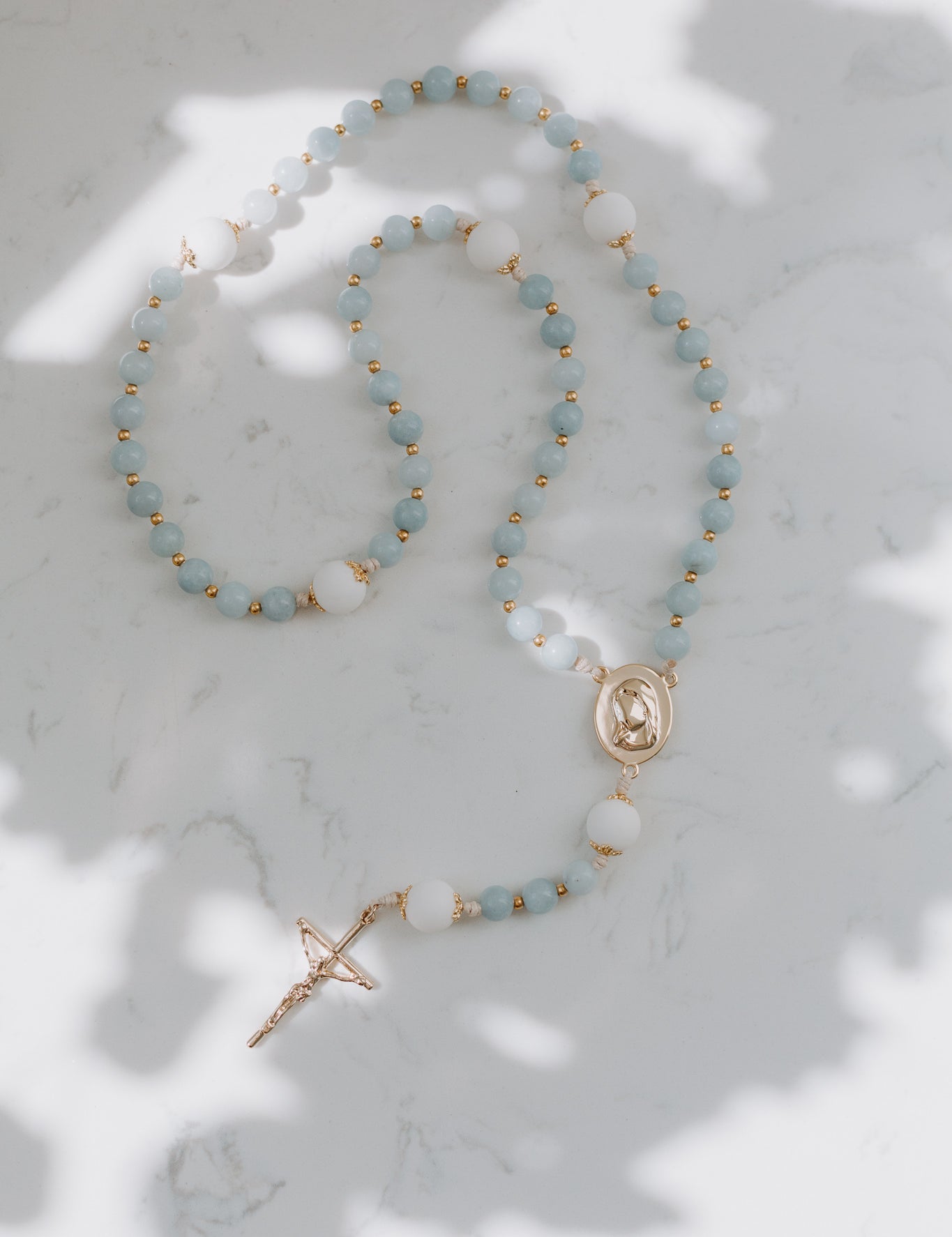 A medjugorje rosary with baby blue and white beads, gold accents, and a gold cross, displayed on a light background.
