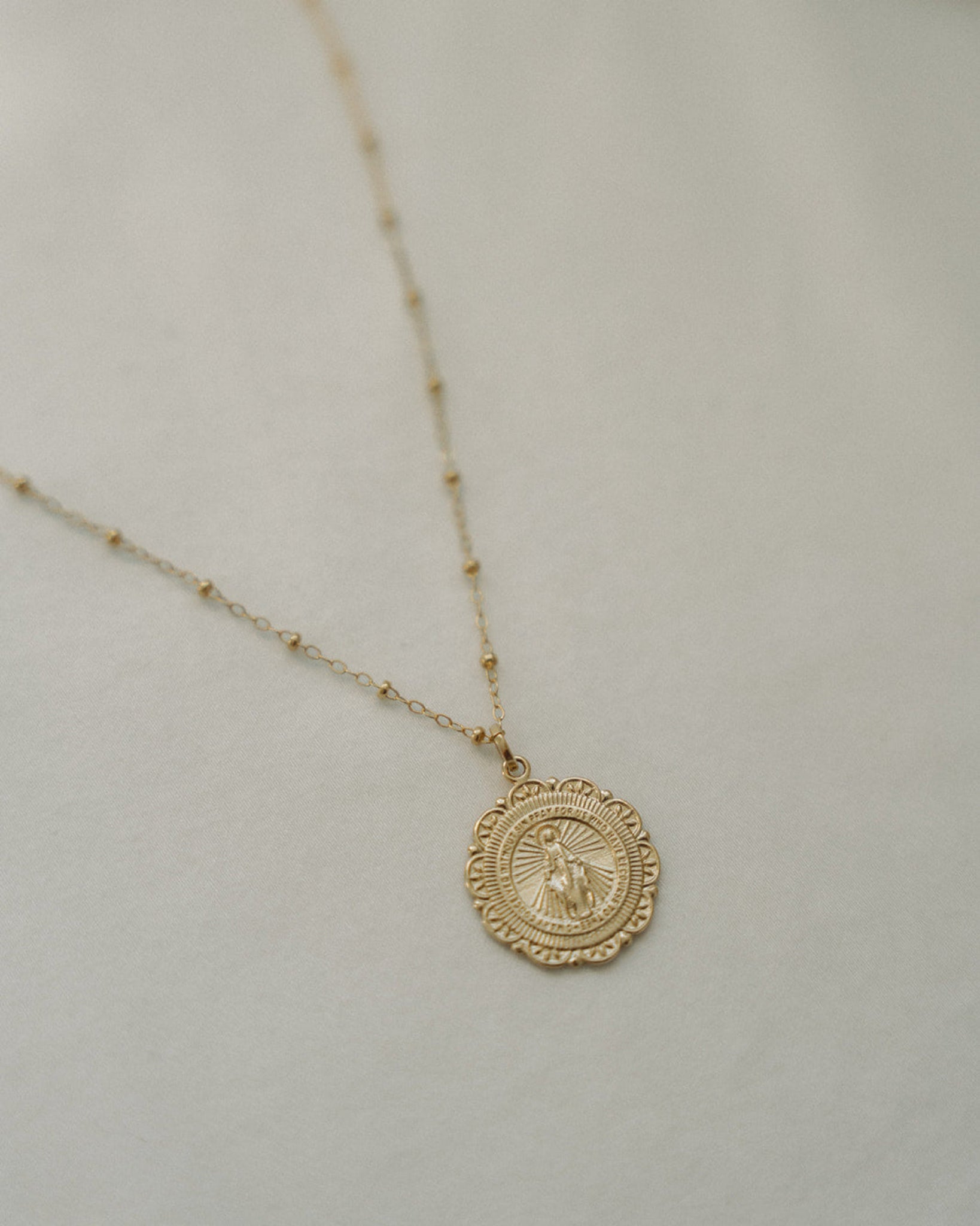 A delicate gold chain necklace featuring a small, detailed circular miraculous medal with a soft, light beige textured background.