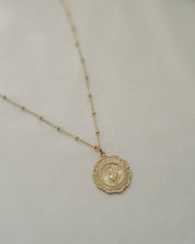 A delicate gold chain necklace featuring a small, detailed circular miraculous medal with a soft, light beige textured background.