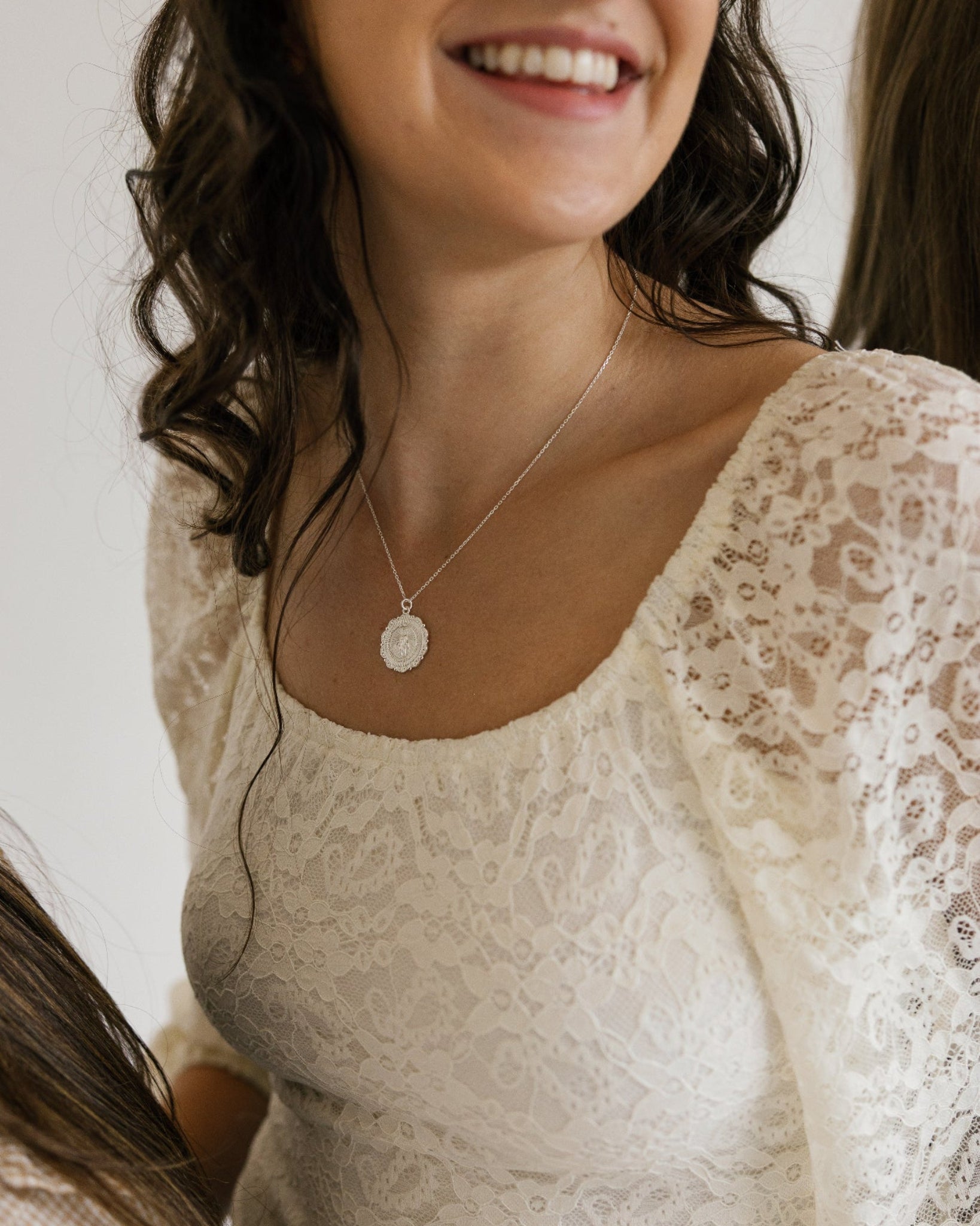A smiling woman wearing a delicate silver miraculous medal necklace paired with a cream lace top. The photo is cropped to show her from the chest up against a plain light background.