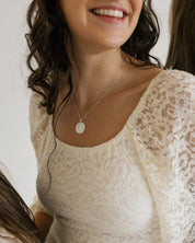 A smiling woman wearing a delicate silver miraculous medal necklace paired with a cream lace top. The photo is cropped to show her from the chest up against a plain light background.