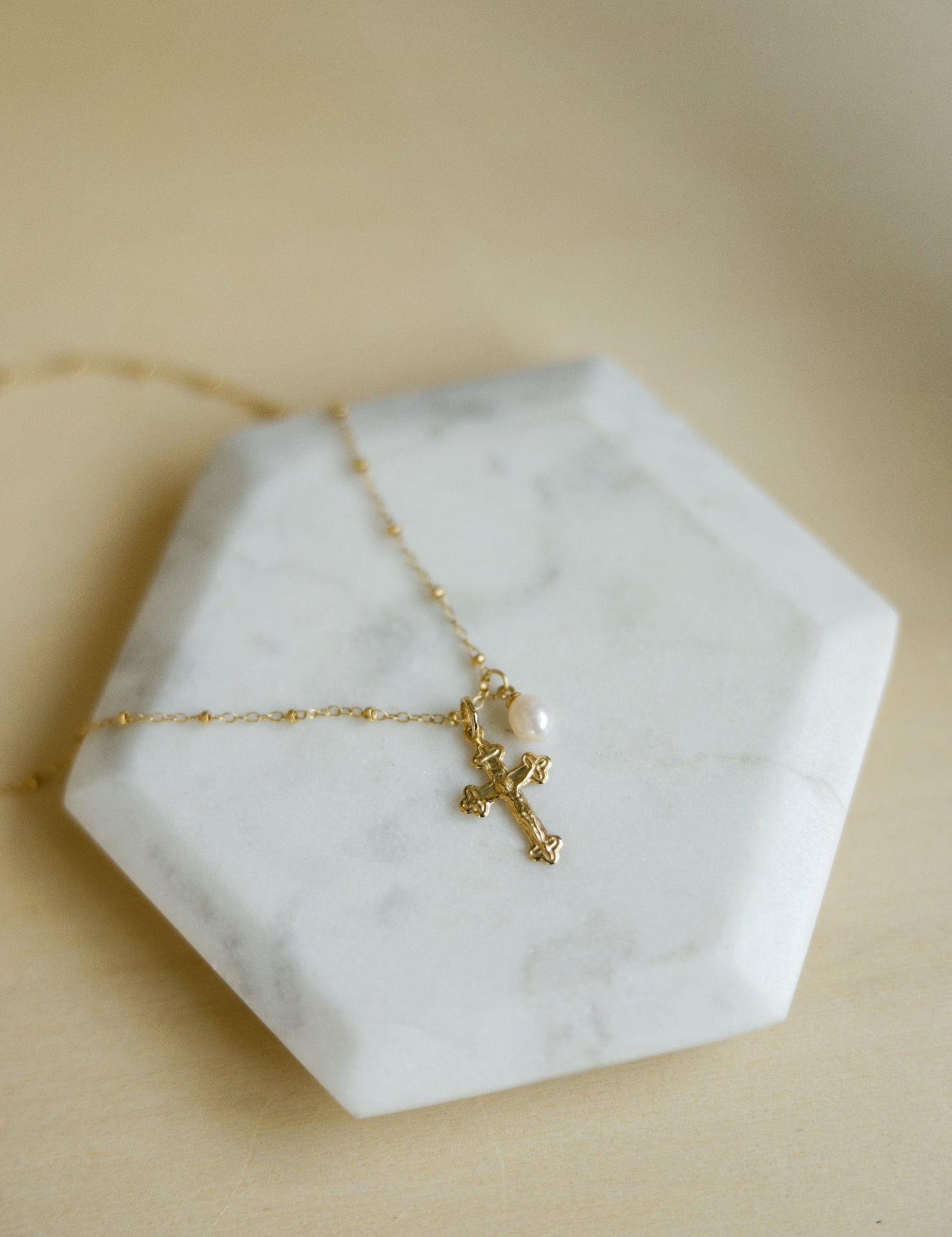 A delicate gold beaded chain necklace with a small gold crucifix and a pearl accent, displayed on a white hexagonal marble tray against a warm beige background.