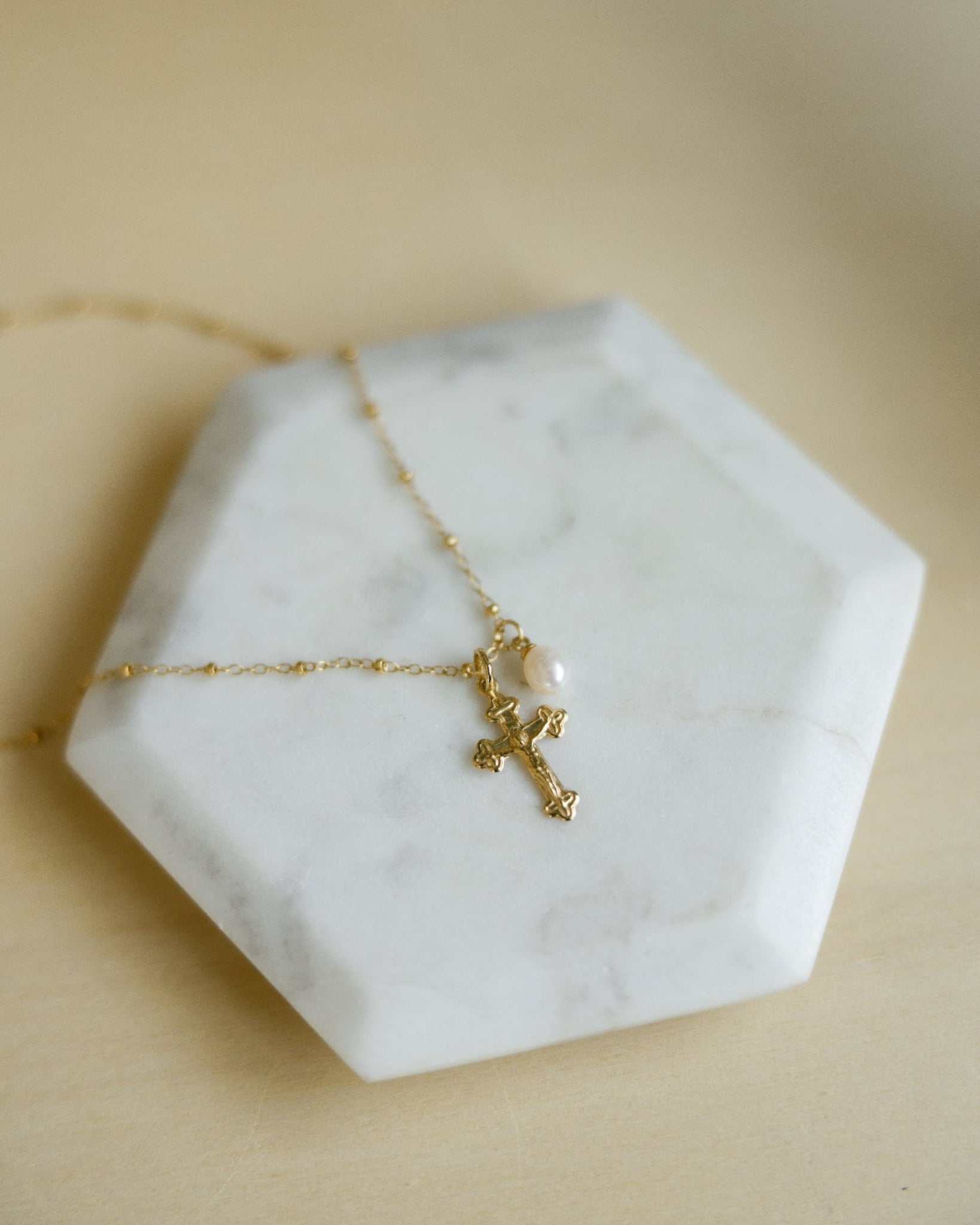 A delicate gold beaded chain necklace with a small gold crucifix and a pearl accent, displayed on a white hexagonal marble tray against a warm beige background.