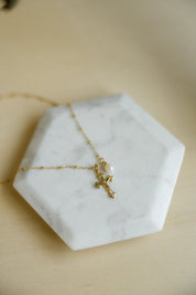 A delicate gold beaded chain necklace with a small gold crucifix and a pearl accent, displayed on a white hexagonal marble tray against a warm beige background.