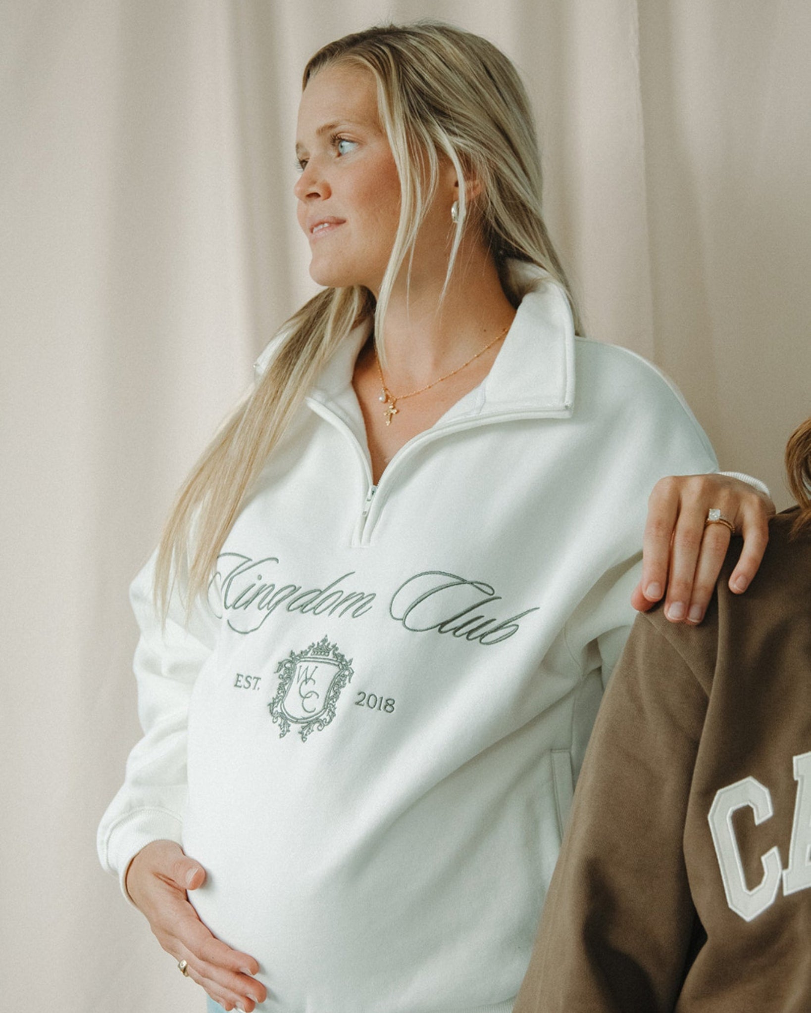 Woman wearing a white sweatshirt with 'Kingdom Club' text, standing next to another person.