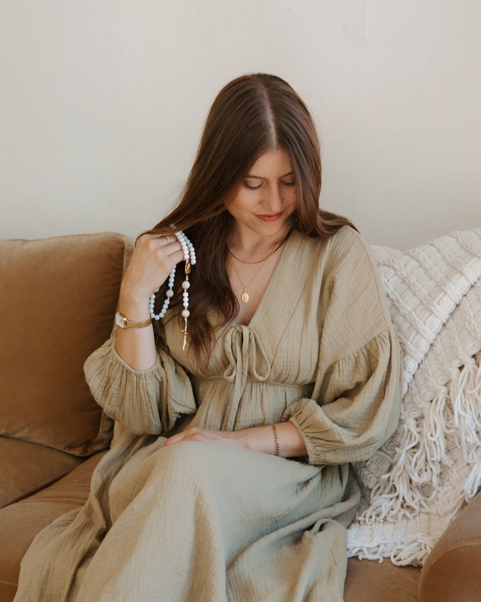 Woman sitting on a couch wearing a beige dress, holding jewelry.