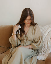 Woman sitting on a couch wearing a beige dress, holding jewelry.