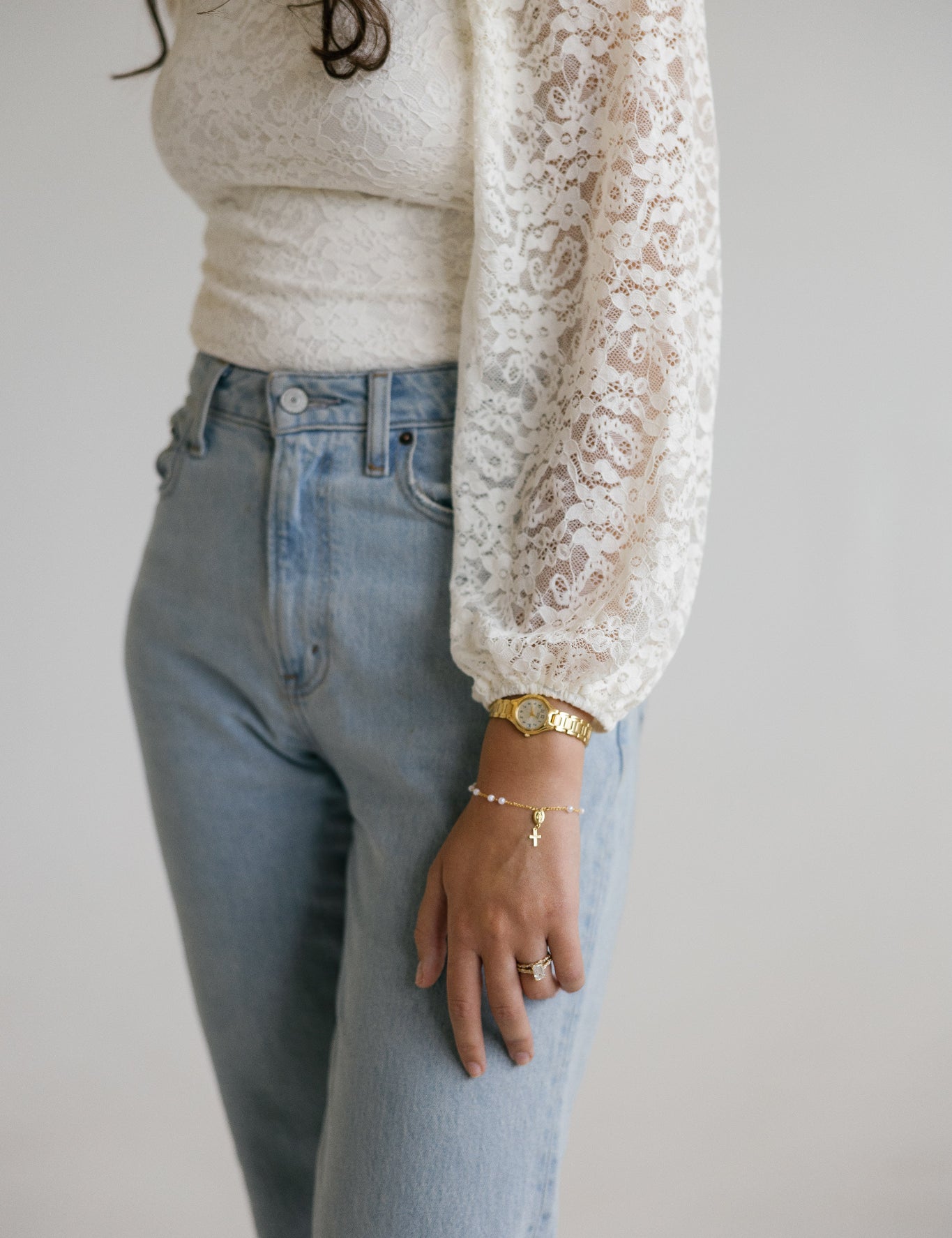 A person wearing a cream lace long-sleeve top over a cream ribbed top and light blue jeans, with gold pearl rosary bracelets, against a plain light gray background.