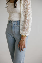 A person wearing a cream lace long-sleeve top over a cream ribbed top and light blue jeans, with gold pearl rosary bracelets, against a plain light gray background.