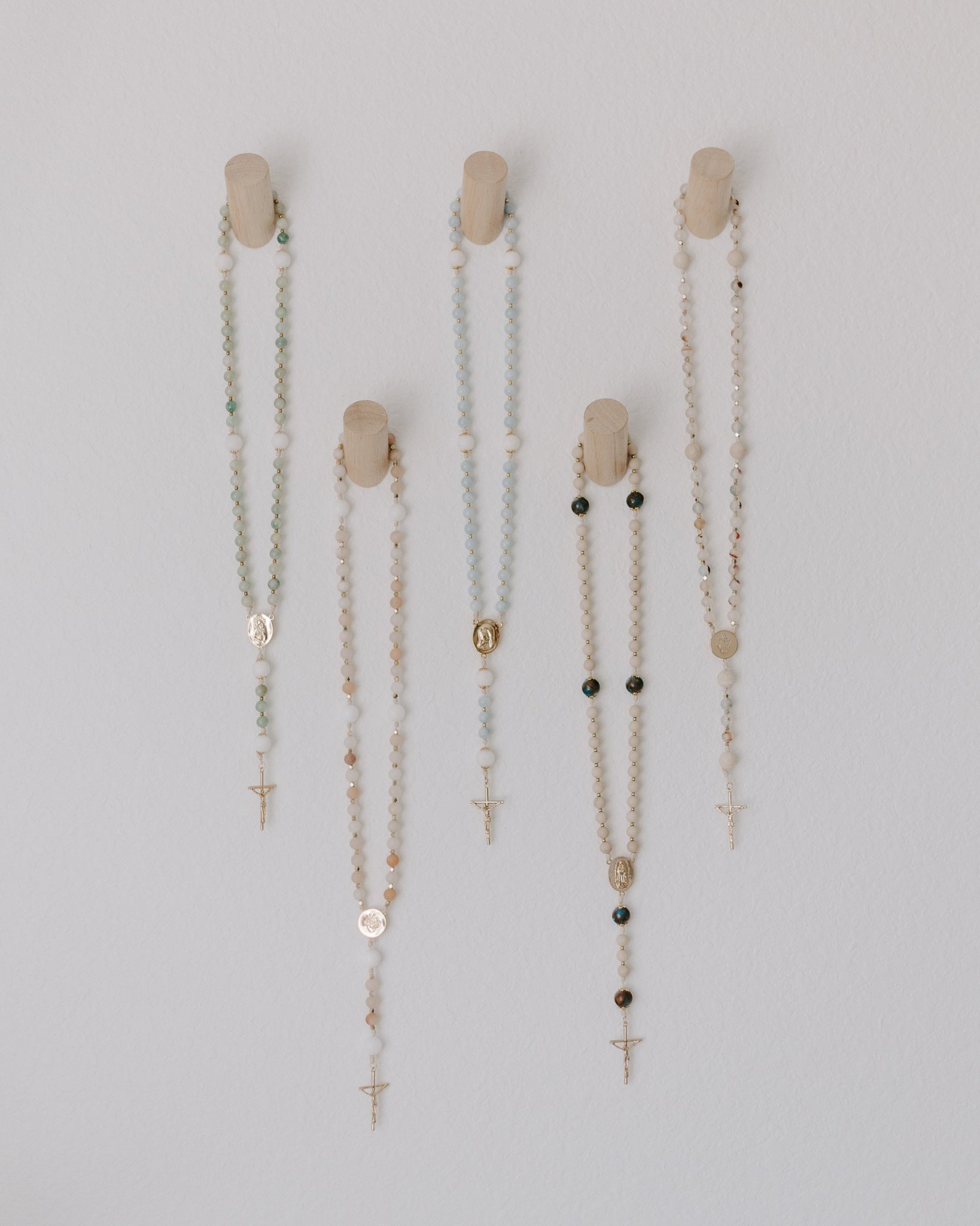 Set of five rosary pegs with rosaries of various bead designs on a light gray background