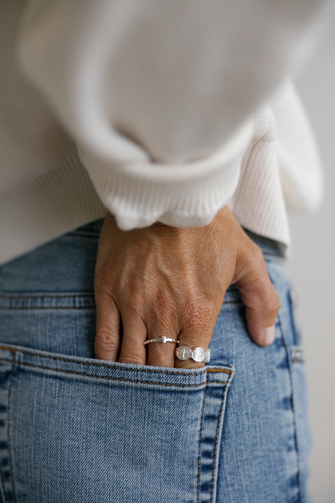 A close-up lifestyle shot of a person wearing silver catholic rings with a white ribbed sweater and blue jeans, looking over their shoulder.