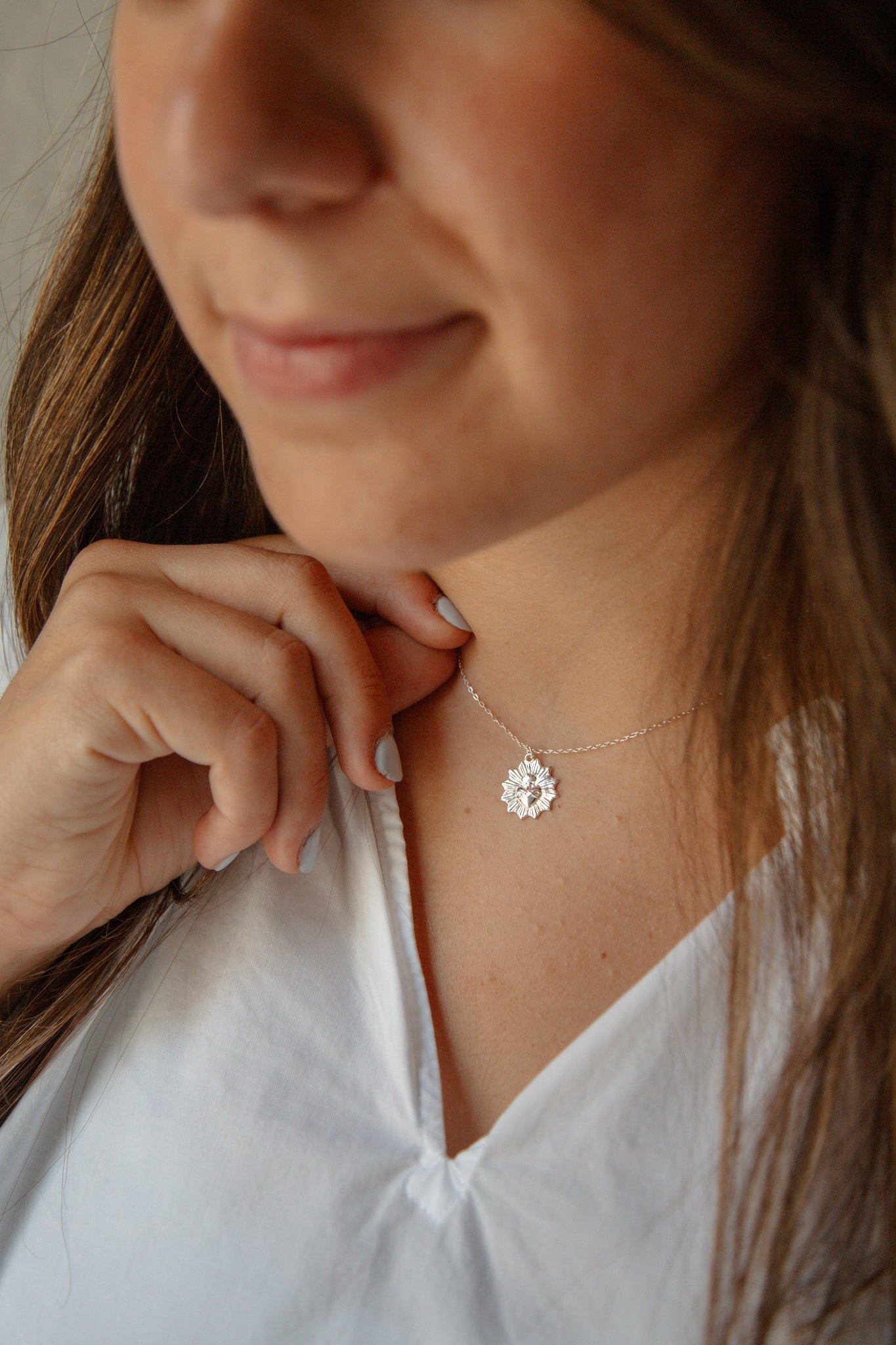 A close-up lifestyle shot of a woman wearing a delicate silver necklace with a small, silver sacred heart medal. She is wearing a white top. 