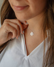 A close-up lifestyle shot of a woman wearing a delicate silver necklace with a small, silver sacred heart medal. She is wearing a white top. 