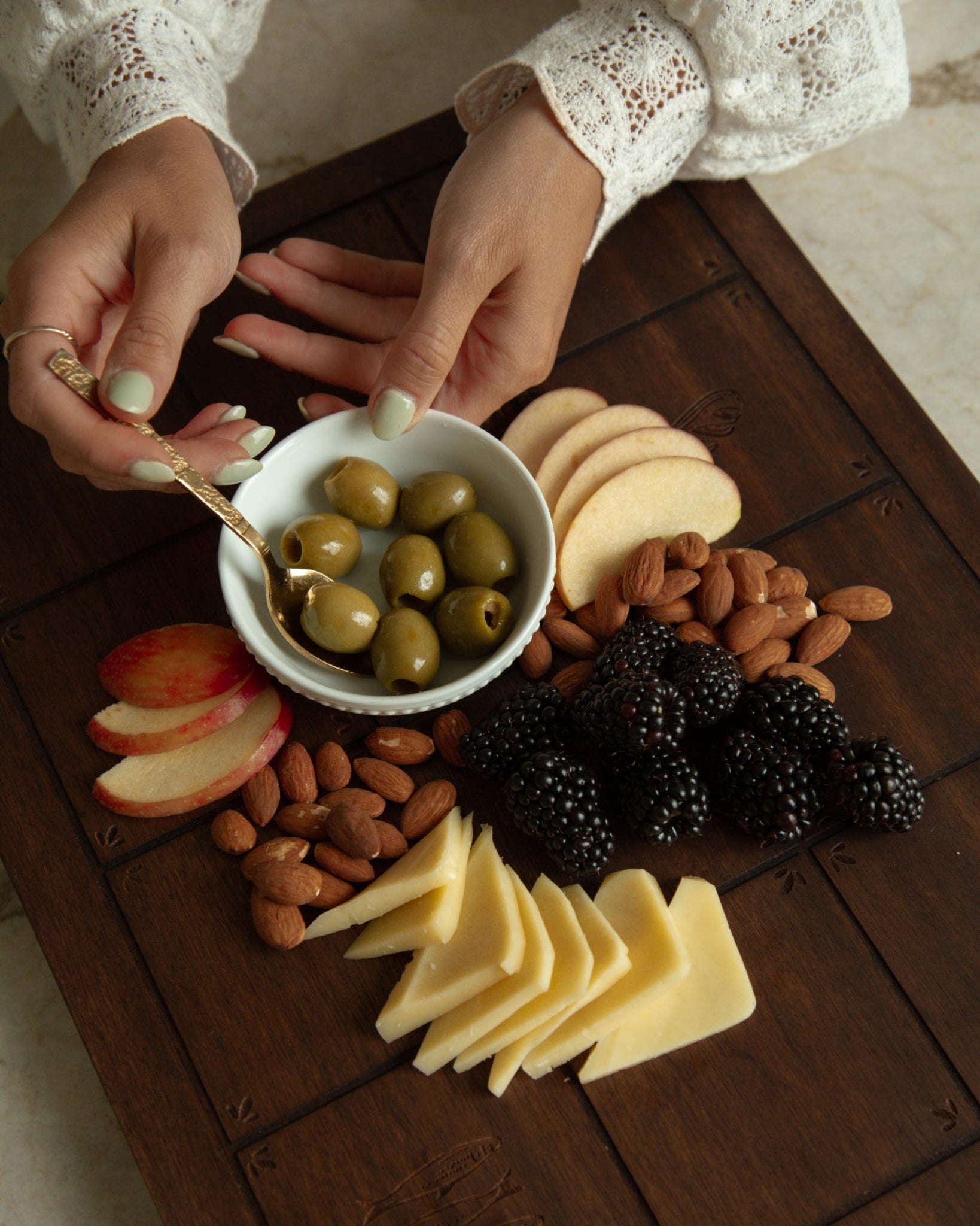 A styled Catholic charcuterie board with sliced cheese, almonds, blackberries, apple slices, and green olives in a small white bowl held by hands wearing a white lace-sleeve top, set on a dark wooden board.